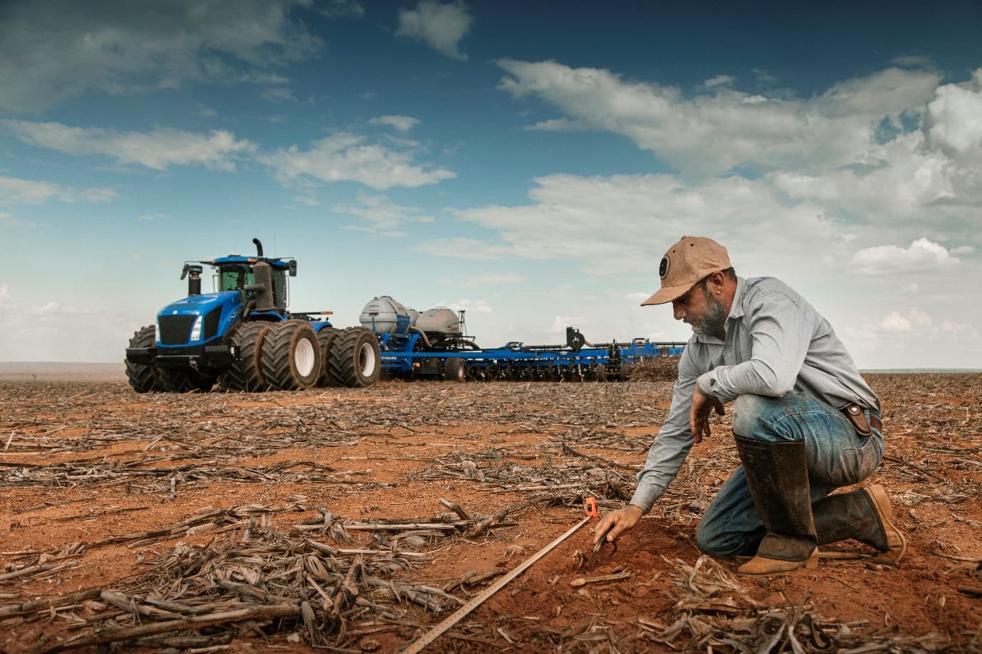 Produtor agachado observando o solo em uma área agrícola, com maquinário ao fundo
